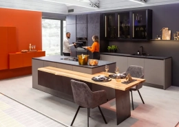Moderne Küche in grau mit schwarzen Arbeitsplatten, schwarzen Armaturen mit Kücheninsel und niedrigem Tresen mit Essplatz. Als Kontrast dazu orange Wand mit gleichfarbigem Sideboard.