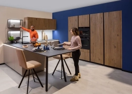 Moderne Küche in L-Form aus einem Materialmix mit Holz, weißen Hochglanzfronten und modernen mattschwarzen Armaturen. Die schwarze Granitarbeitsplatte geht in einen Essplatz über. Kühlschrank, Backofen und Vorratsschränke sind in die Wand integriert.